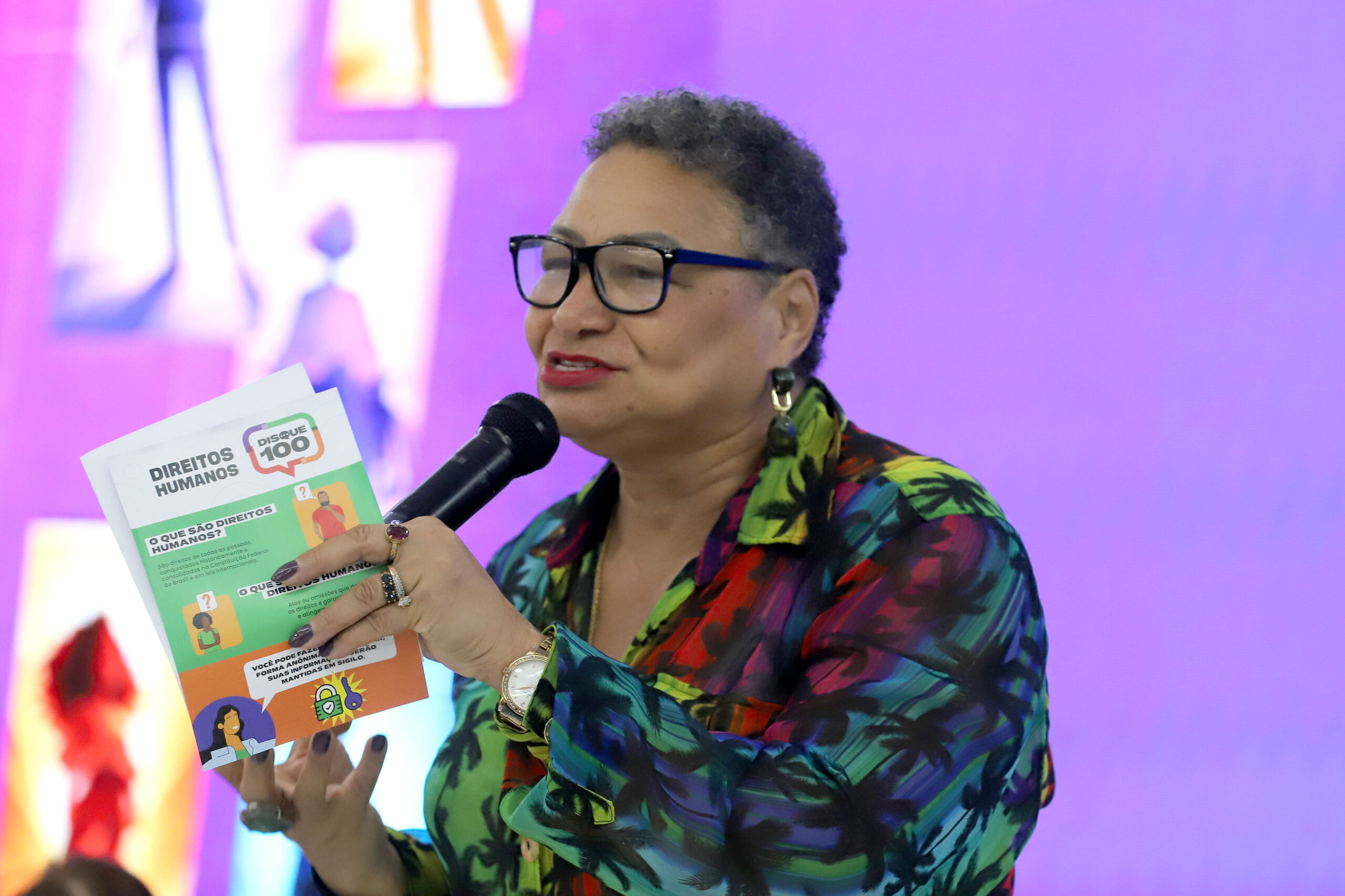 Encontro Nacional fortalece protagonismo de mulheres com deficiência e impulsiona agenda de inclusão no país Mulher parda, de cabelo curto grisalho e óculos, segura cartilha sobre direitos humanos enquanto fala ao microfone. Usa blusa estampada colorida. Fundo com painel iluminado.