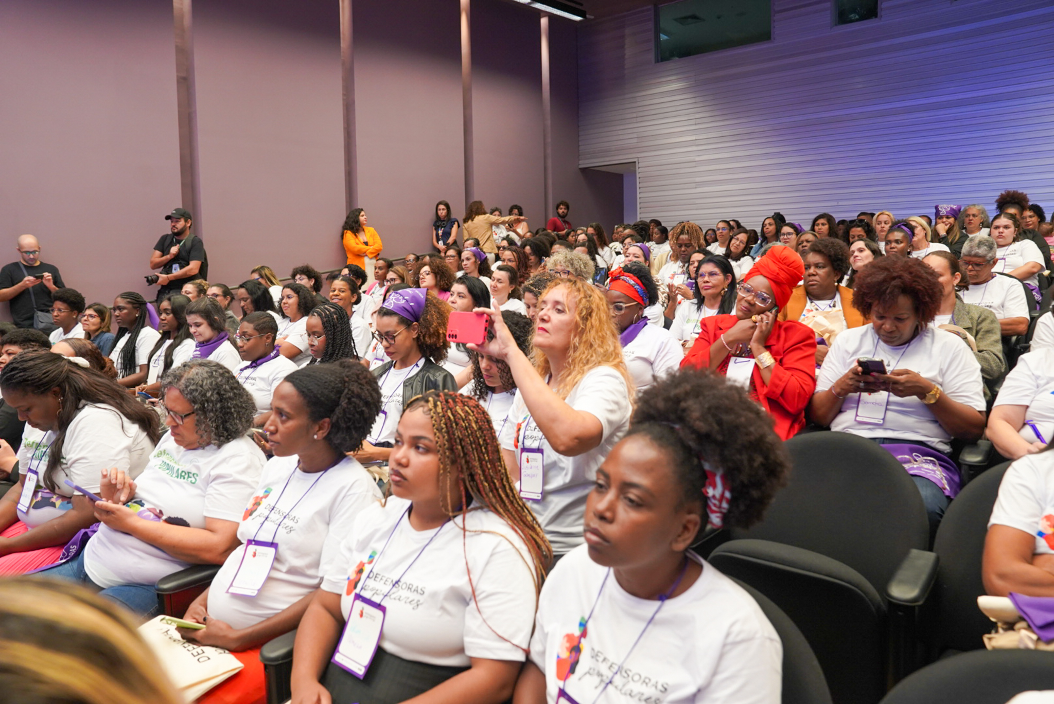 MDHC firma parceria para preservação da memória, fortalece defensoras populares e reforça a promoção da liberdade religiosa em Minas Gerais Auditório lotado com fileiras de cadeiras pretas. Mulheres usam camisetas brancas e crachás, algumas mexem no celular e outras observam. Ao centro, uma pessoa levanta o celular para registrar o momento.