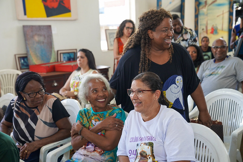 MDHC firma parceria para preservação da memória, fortalece defensoras populares e reforça a promoção da liberdade religiosa em Minas Gerais Sala com cadeiras brancas e quadros nas paredes. Mulheres de diferentes idades conversam e sorriem. Ao centro, a ministra Macaé Evaristo está em pé, interagindo com três mulheres sentadas, em clima acolhedor.