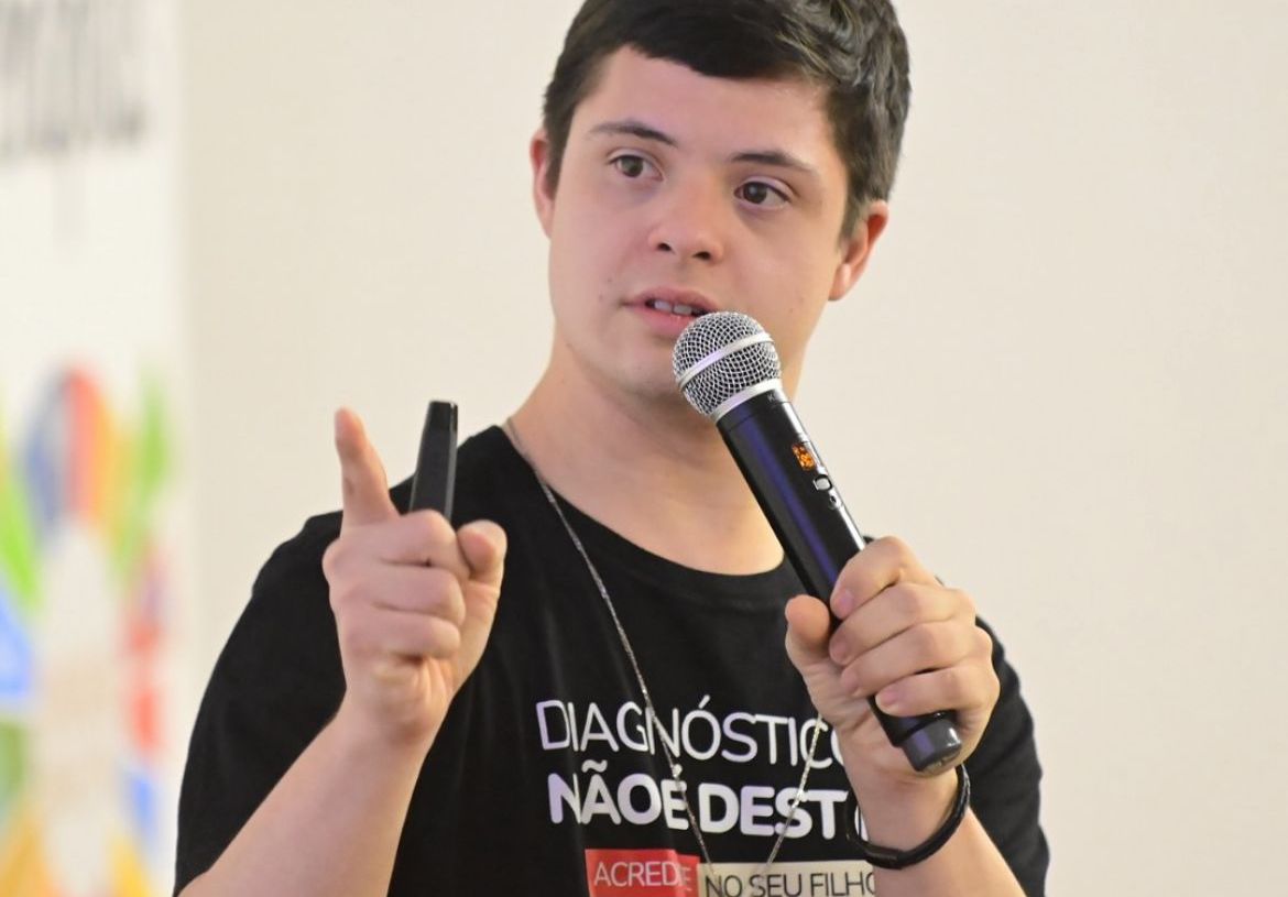Jovem com síndrome de Down segura um microfone e fala em público. Ele veste camiseta preta com a frase “Diagnóstico não é destino” e aponta com o dedo enquanto se expressa. O fundo é claro e desfocado.