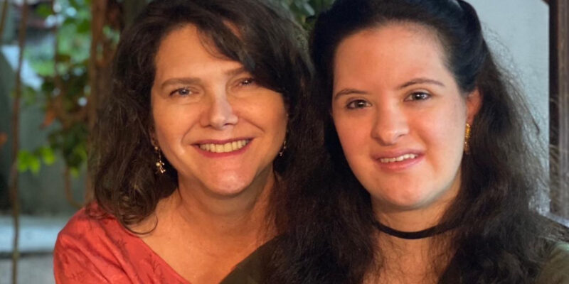 Ana Figueiredo e Jéssica Mendes de Figueiredo aparecem sentadas lado a lado, sorrindo para a câmera. Jéssica, jovem com síndrome de Down, tem cabelos longos e veste blusa verde com colar preto. Ana usa blusa vermelha e brincos. Ao fundo, plantas e iluminação suave em ambiente interno.