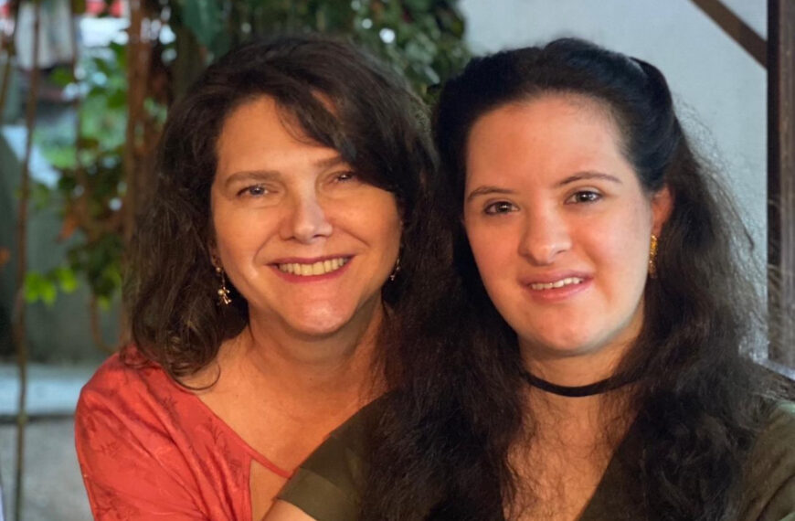 Ana Figueiredo e Jéssica Mendes de Figueiredo aparecem sentadas lado a lado, sorrindo para a câmera. Jéssica, jovem com síndrome de Down, tem cabelos longos e veste blusa verde com colar preto. Ana usa blusa vermelha e brincos. Ao fundo, plantas e iluminação suave em ambiente interno.