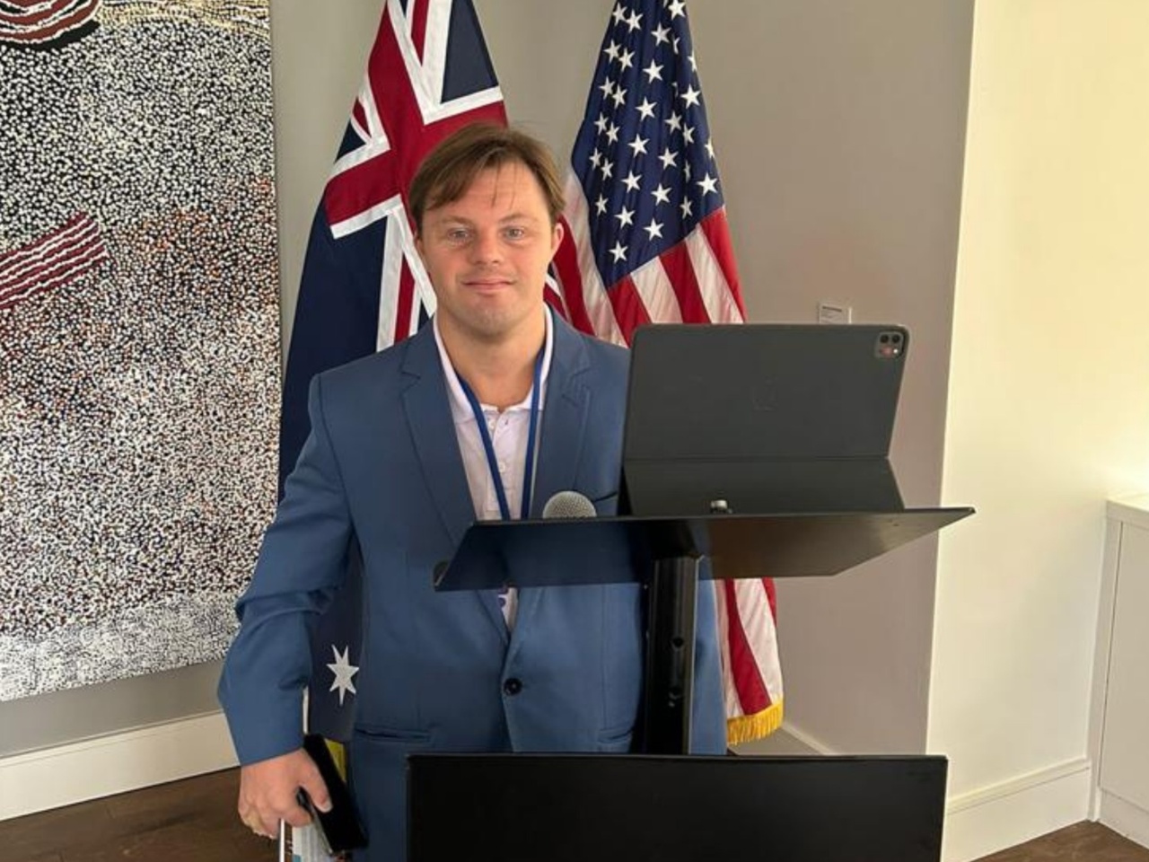 Homem com síndrome de Down está em pé atrás de um púlpito, em ambiente interno, como em uma apresentação. Ele veste blazer azul e camisa clara, com crachá no pescoço. À sua frente há um tablet e um microfone. Ao fundo aparecem bandeiras dos Estados Unidos e da Austrália, além de um painel artístico na parede.