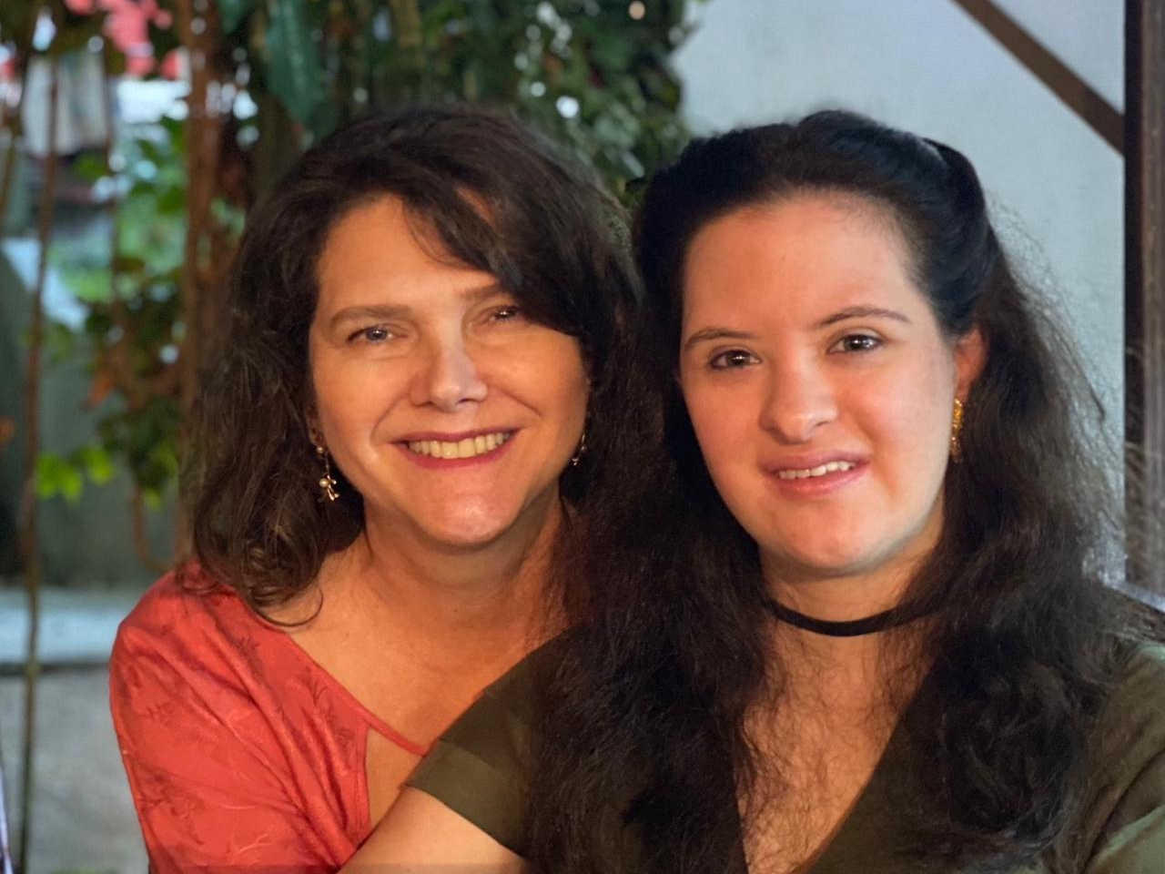 Ana Figueiredo e Jéssica Mendes de Figueiredo aparecem sentadas lado a lado, sorrindo para a câmera. Jéssica, jovem com síndrome de Down, tem cabelos longos e veste blusa verde com colar preto. Ana usa blusa vermelha e brincos. Ao fundo, plantas e iluminação suave em ambiente interno.