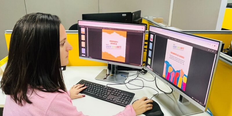 Mulher trabalha em computador com dois monitores em um escritório. Nas telas aparecem documentos do plano Novo Viver sem Limite, incluindo a capa “Orientação à Adesão ao Novo Viver sem Limite” e um documento de suporte. Sobre a mesa há teclado, mouse e telefone.