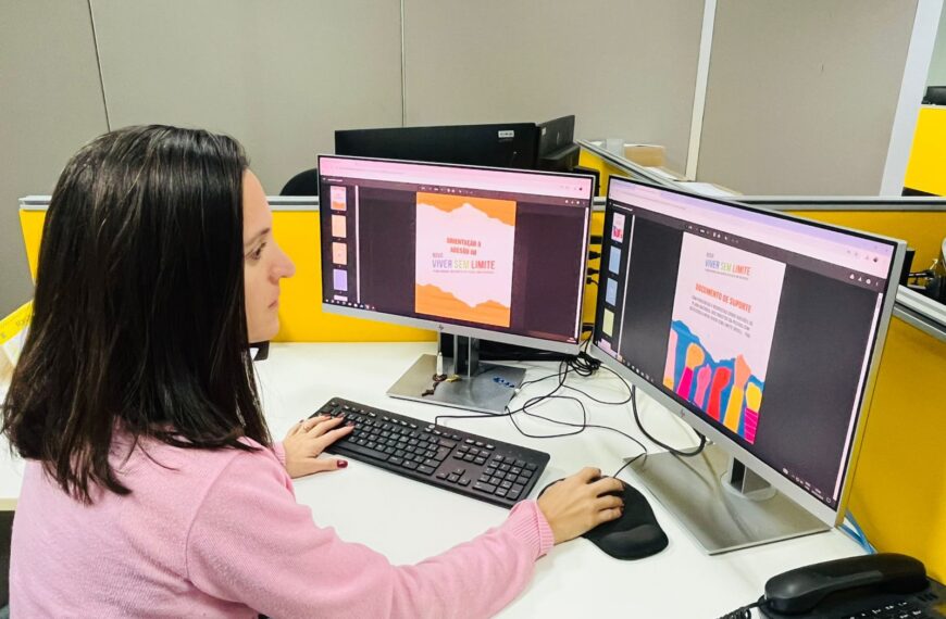 Mulher trabalha em computador com dois monitores em um escritório. Nas telas aparecem documentos do plano Novo Viver sem Limite, incluindo a capa “Orientação à Adesão ao Novo Viver sem Limite” e um documento de suporte. Sobre a mesa há teclado, mouse e telefone.