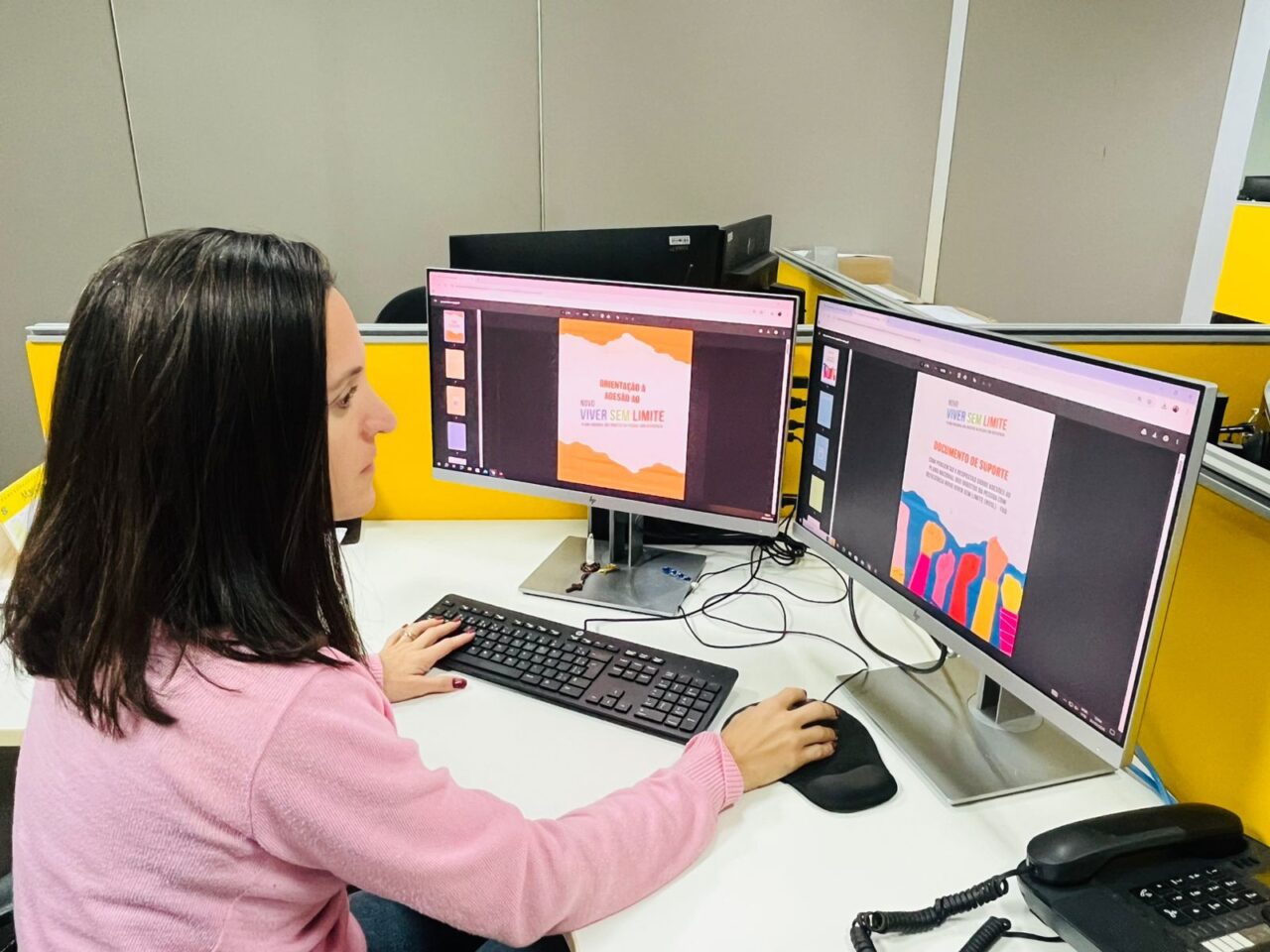 Mulher trabalha em computador com dois monitores em um escritório. Nas telas aparecem documentos do plano Novo Viver sem Limite, incluindo a capa “Orientação à Adesão ao Novo Viver sem Limite” e um documento de suporte. Sobre a mesa há teclado, mouse e telefone.