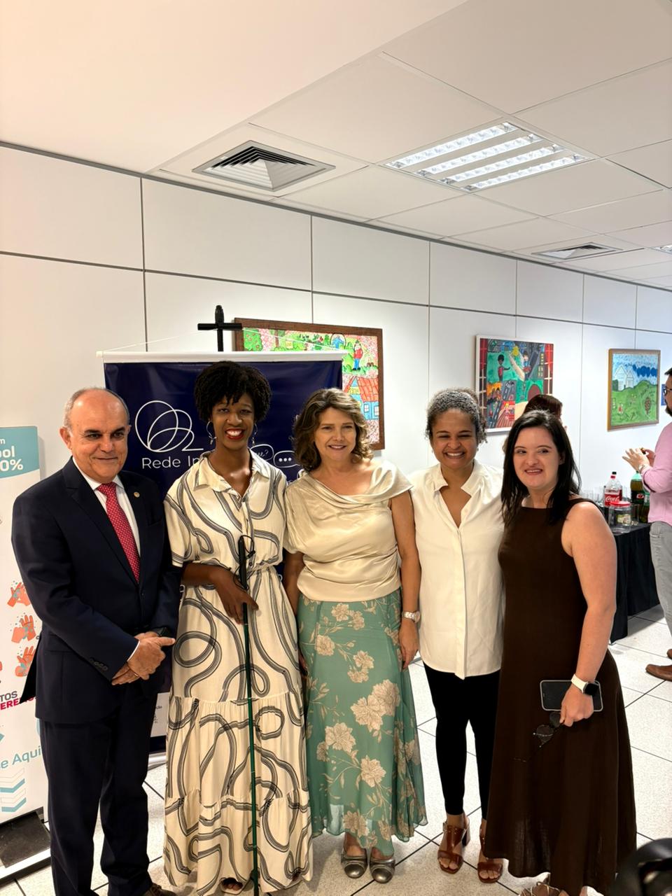 Um homem e quatro mulheres posam lado a lado em espaço interno de evento. Ao fundo há banner da Rede In e quadros coloridos na parede. O grupo sorri para a foto; uma das mulheres utiliza bengala verde.