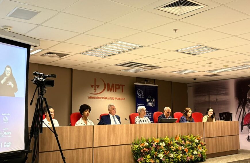 Mesa de autoridades em auditório do Ministério Público do Trabalho (MPT) durante seminário. Sete participantes estão sentados em painel, com intérprete de Libras projetada em telão ao lado e arranjo de flores à frente.