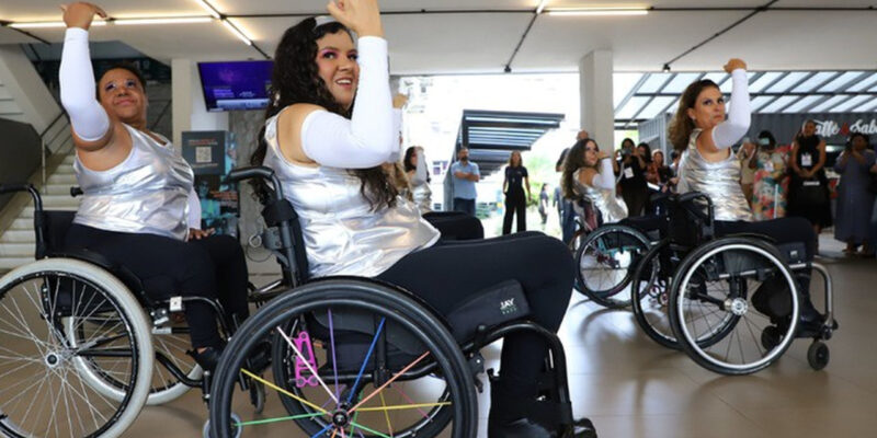Três mulheres em cadeiras de rodas realizam uma apresentação de dança em um espaço interno iluminado. Vestem blusas prateadas e calças pretas, com braços erguidos na coreografia. Ao fundo, público observa e registra o momento.