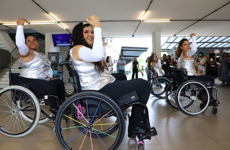Três mulheres em cadeiras de rodas realizam uma apresentação de dança em um espaço interno iluminado. Vestem blusas prateadas e calças pretas, com braços erguidos na coreografia. Ao fundo, público observa e registra o momento.