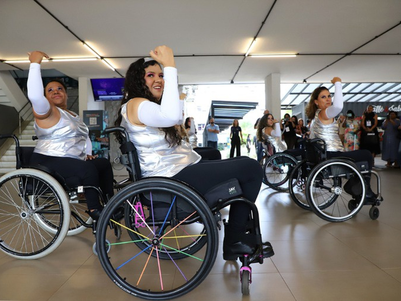 Três mulheres em cadeiras de rodas realizam uma apresentação de dança em um espaço interno iluminado. Vestem blusas prateadas e calças pretas, com braços erguidos na coreografia. Ao fundo, público observa e registra o momento.