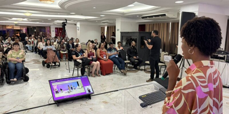 Mulher negra, Isadora Nascimento, aparece em primeiro plano, de perfil, falando ao microfone atrás de um púlpito transparente. À frente, público diverso acompanha a atividade, incluindo pessoas com deficiência.