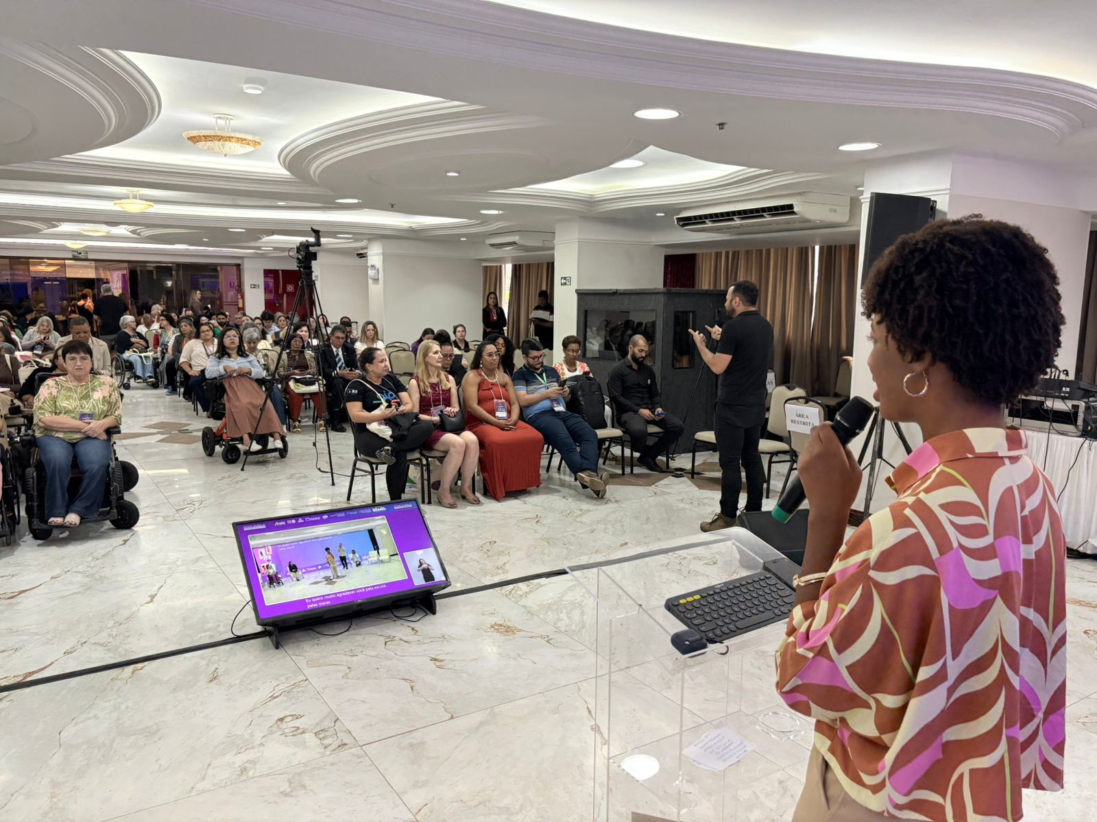 Mulher negra, Isadora Nascimento, aparece em primeiro plano, de perfil, falando ao microfone atrás de um púlpito transparente. À frente, público diverso acompanha a atividade, incluindo pessoas com deficiência.