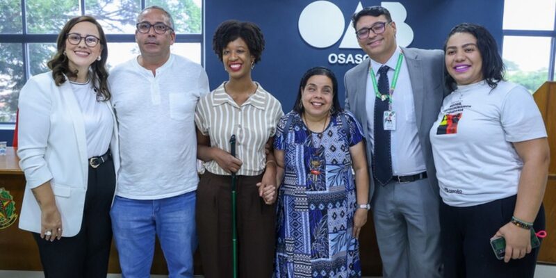 Grupo de homens e mulheres posa lado a lado em ambiente institucional, diante de parede azul com a palavra “Osasco”. Entre eles, mulher com bengala e mulher com nanismo. Todos sorriem para a câmera em registro oficial.