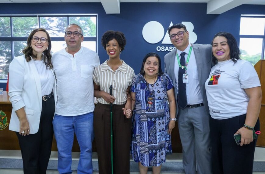 Grupo de homens e mulheres posa lado a lado em ambiente institucional, diante de parede azul com a palavra “Osasco”. Entre eles, mulher com bengala e mulher com nanismo. Todos sorriem para a câmera em registro oficial.