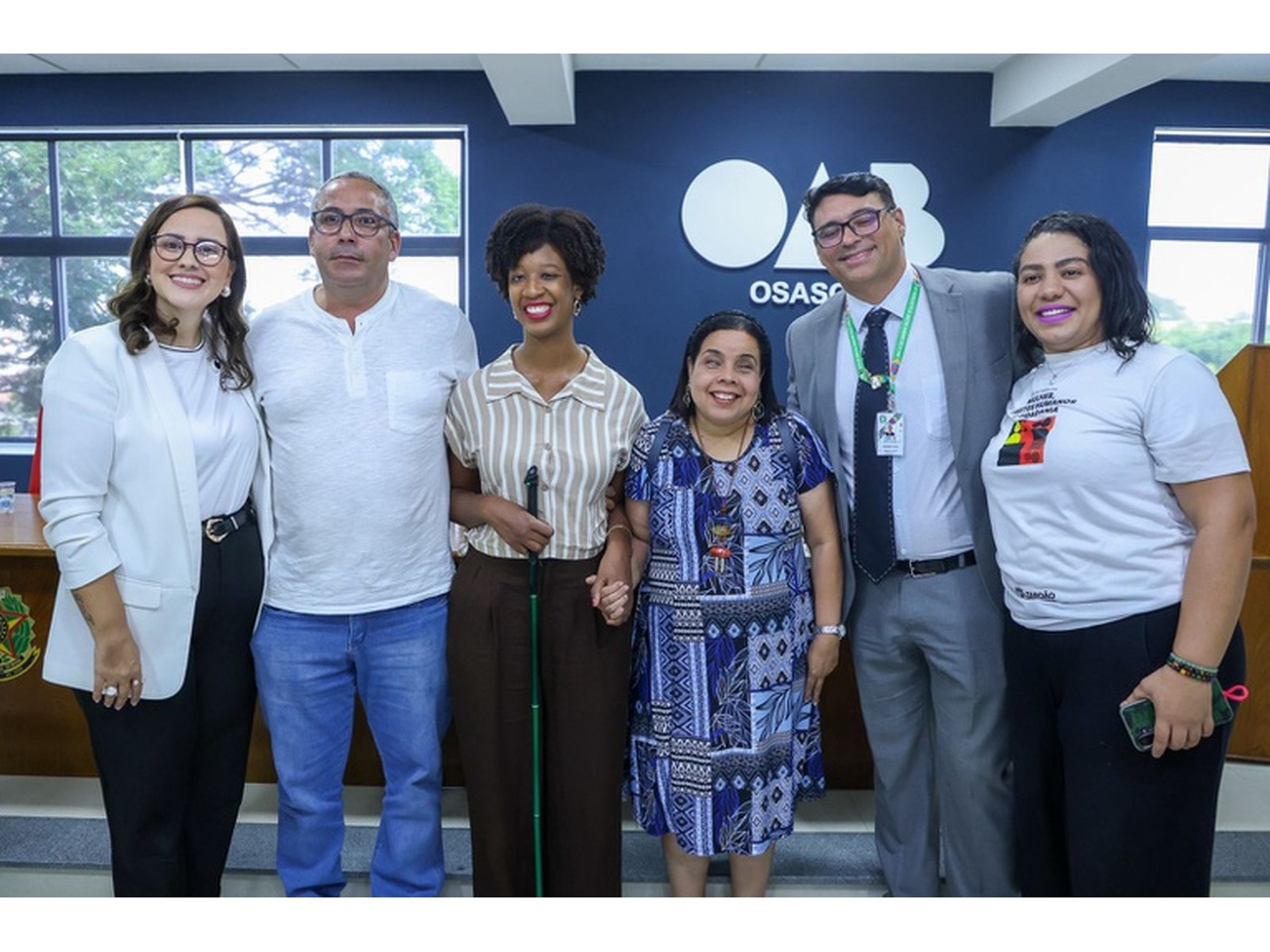 Grupo de homens e mulheres posa lado a lado em ambiente institucional, diante de parede azul com a palavra “Osasco”. Entre eles, mulher com bengala e mulher com nanismo. Todos sorriem para a câmera em registro oficial.