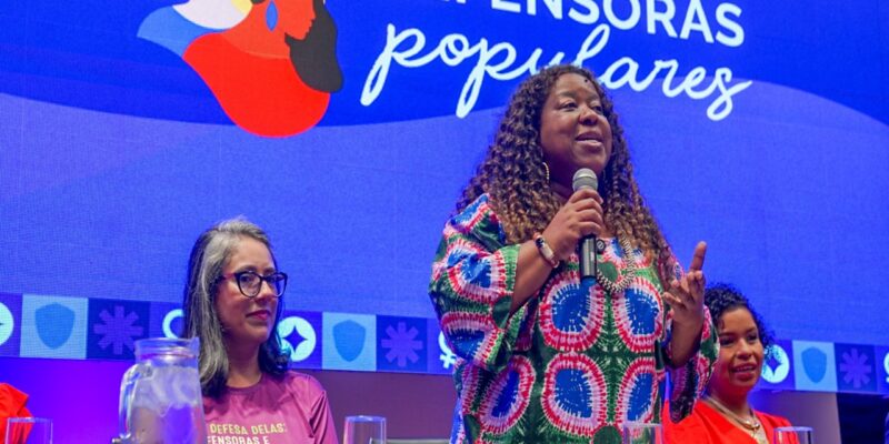 Palco iluminado em azul e roxo, com painel “Defensoras Populares” ao fundo. À mesa, três mulheres sentadas. Ao centro, a ministra Macaé Evaristo fala ao microfone, olhando à frente.
