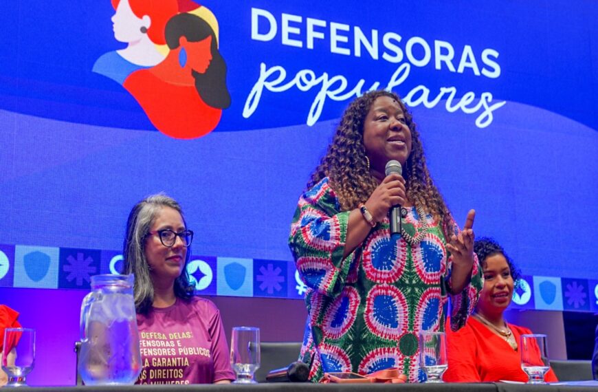 Palco iluminado em azul e roxo, com painel “Defensoras Populares” ao fundo. À mesa, três mulheres sentadas. Ao centro, a ministra Macaé Evaristo fala ao microfone, olhando à frente.