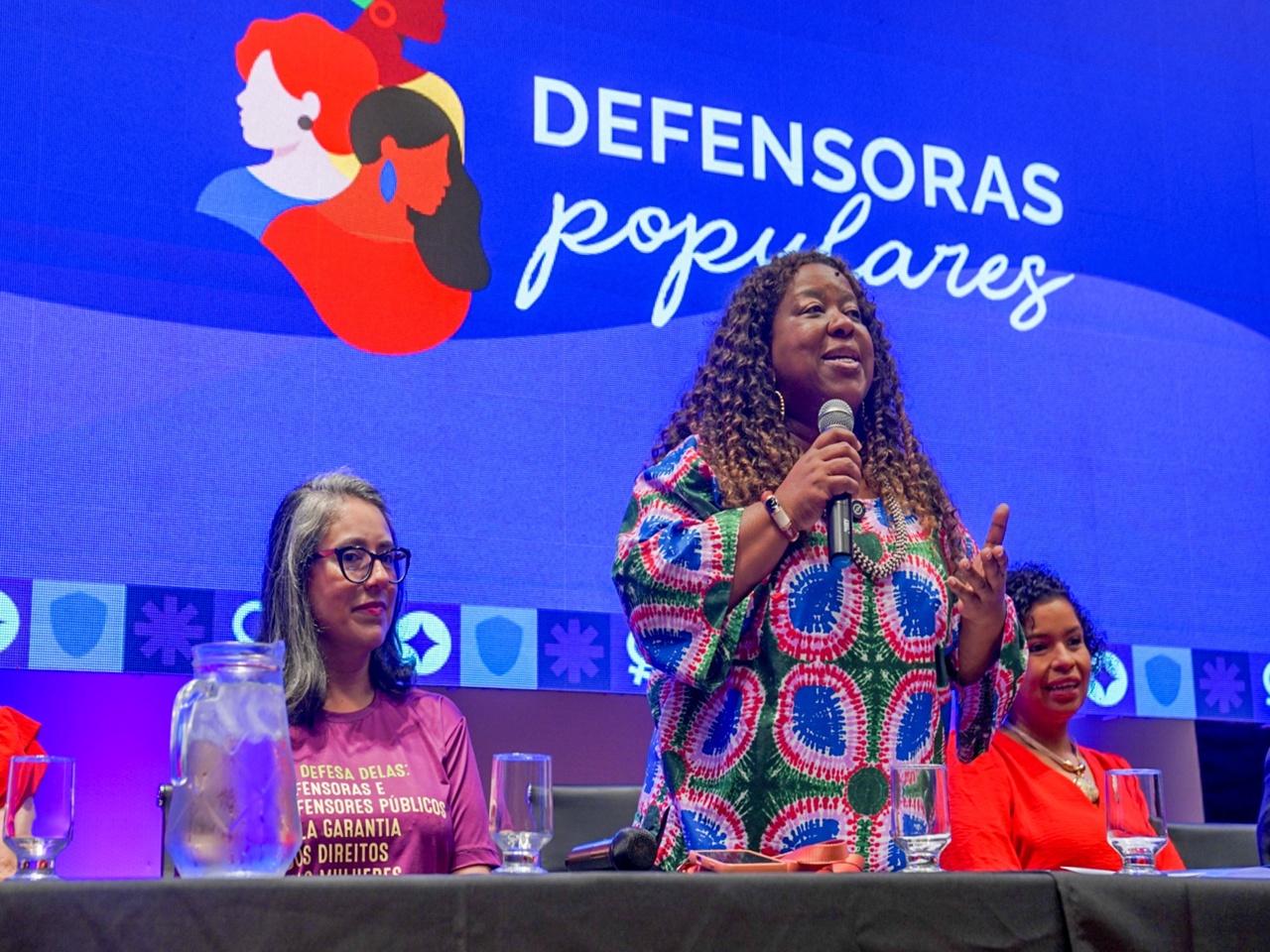Palco iluminado em azul e roxo, com painel “Defensoras Populares” ao fundo. À mesa, três mulheres sentadas. Ao centro, a ministra Macaé Evaristo fala ao microfone, olhando à frente.