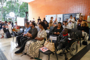 Público acompanha painel no FomentaCine, com pessoas sentadas em fileiras, incluindo pessoa em cadeira de rodas. Há cadeiras reservadas, equipe de filmagem, iluminação e participantes usando celulares.