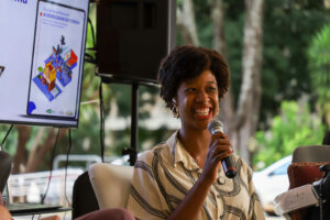 Isadora Nascimento, mulher negra, sorri enquanto fala ao microfone em painel sobre acessibilidade no cinema. Ao lado, tela exibe guia com QR code; ao fundo, área externa com árvores.