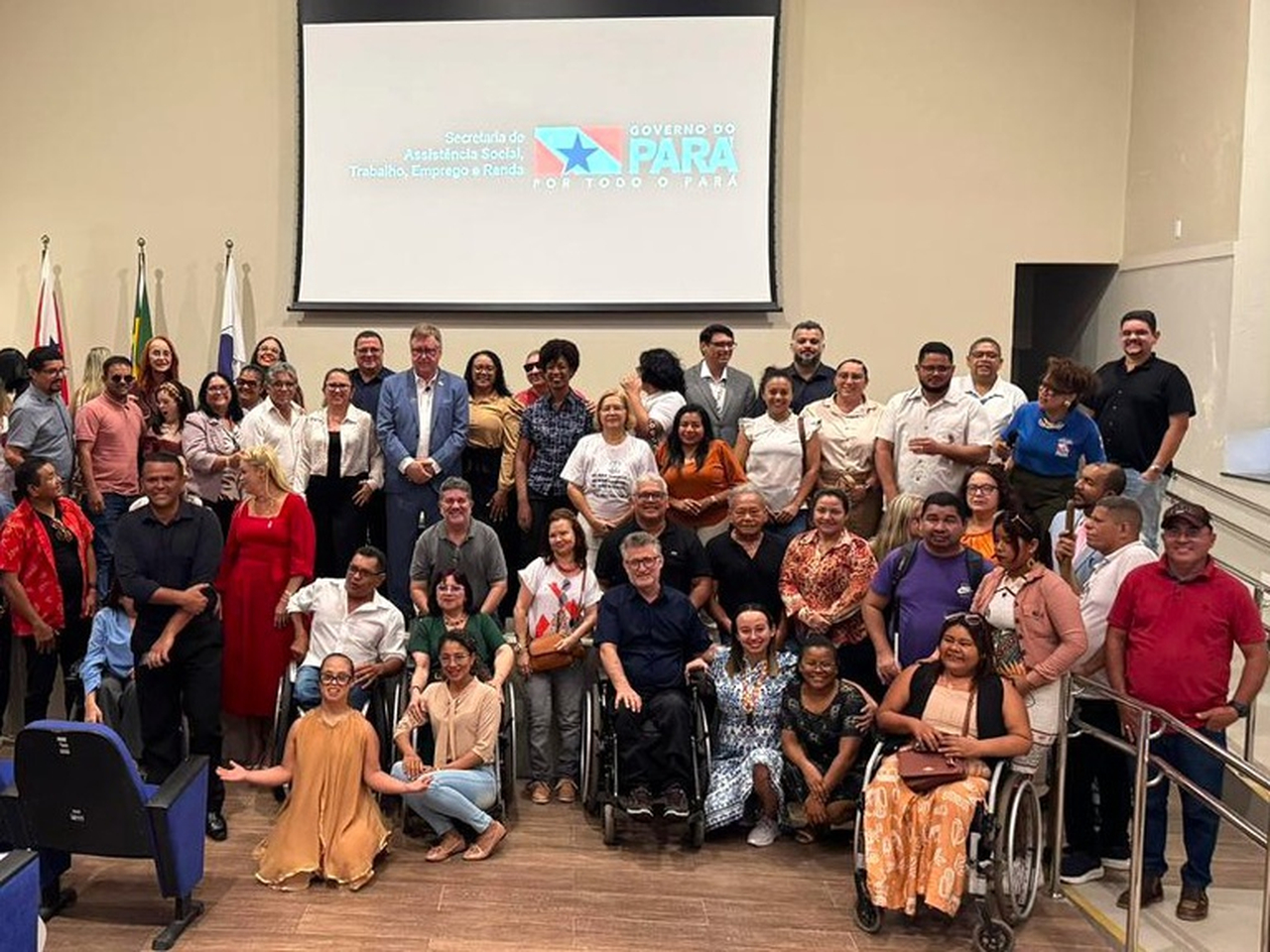 Grupo de homens e mulheres posa para foto em auditório, incluindo pessoas com deficiência, como usuários de cadeira de rodas. Ao fundo, tela com logomarca do Governo do Pará e bandeiras.