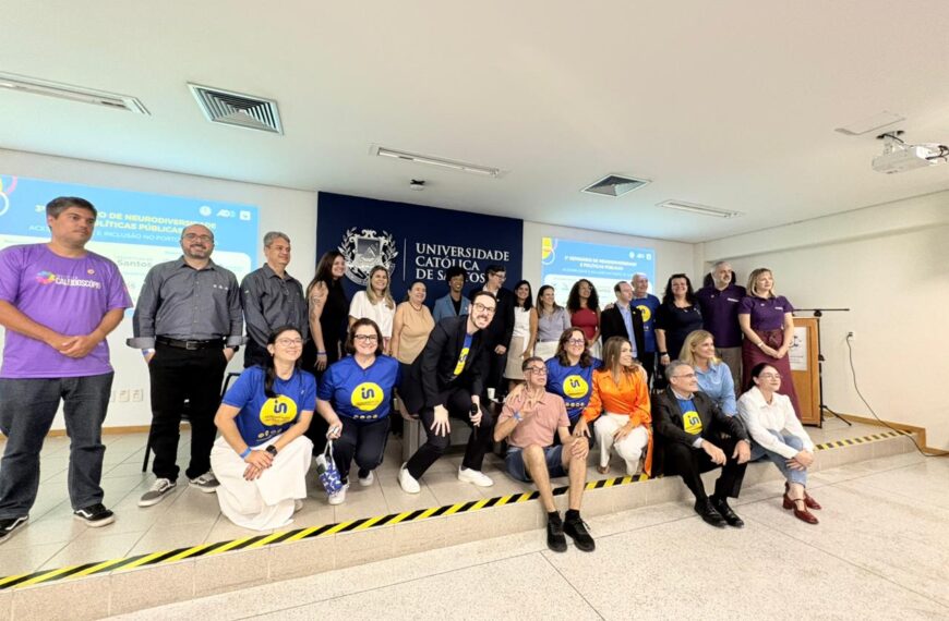 Grupo de cerca de 20 pessoas, entre homens e mulheres, posa em um auditório universitário, em frente ao painel da Universidade Católica de Santos. Parte está em pé e parte sentada, com algumas pessoas usando camisetas azuis com símbolo amarelo de inclusão. Ao fundo, há projeções sobre neurodiversidade e políticas públicas.