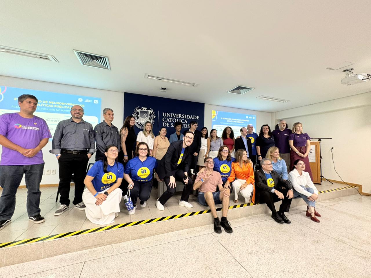 Grupo de cerca de 20 pessoas, entre homens e mulheres, posa em um auditório universitário, em frente ao painel da Universidade Católica de Santos. Parte está em pé e parte sentada, com algumas pessoas usando camisetas azuis com símbolo amarelo de inclusão. Ao fundo, há projeções sobre neurodiversidade e políticas públicas.