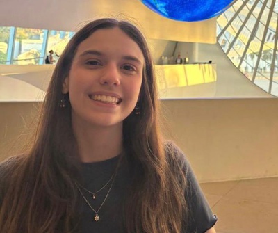 Mulher jovem de cabelo longo e castanho sorri para a câmera em ambiente interno. Ao fundo, há paredes em tom bege, janelas grandes e uma grande esfera azul suspensa no teto.