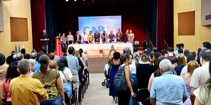 Auditório cheio, com público em pé voltado para o palco. À frente, autoridades formam uma mesa solene sob luzes e cortinas vermelhas. Uma pessoa registra o momento com o celular no corredor central.
