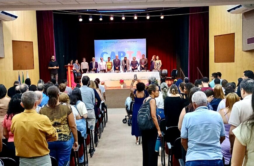 Auditório cheio, com público em pé voltado para o palco. À frente, autoridades formam uma mesa solene sob luzes e cortinas vermelhas. Uma pessoa registra o momento com o celular no corredor central.