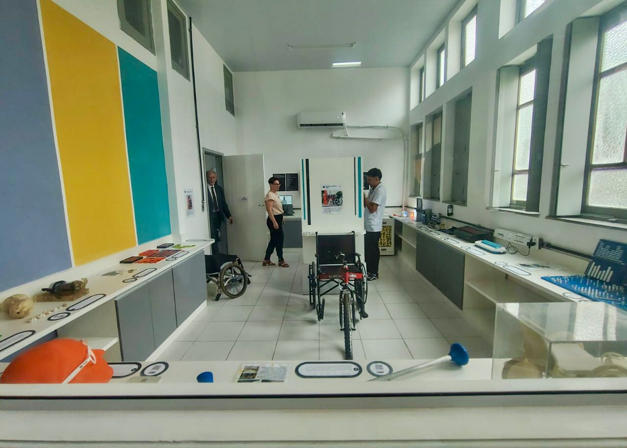 Governo do Brasil e Instituto Benjamin Constant ampliam acesso à tecnologia assistiva e marcam nova etapa do Novo Viver sem Limite Sala com janelas altas à direita, por onde entra luz natural, e mesas distribuídas nas laterais exibindo recursos de tecnologia assistiva, como próteses, adaptações para mãos, materiais táteis, lupas, peças educativas e dispositivos de apoio. Ao centro, há uma cadeira de rodas adaptada posicionada em destaque. Três pessoas circulam pelo ambiente: um homem observa itens em uma bancada, enquanto uma mulher e outro homem entram por uma porta ao fundo. As paredes têm faixas verticais coloridas.