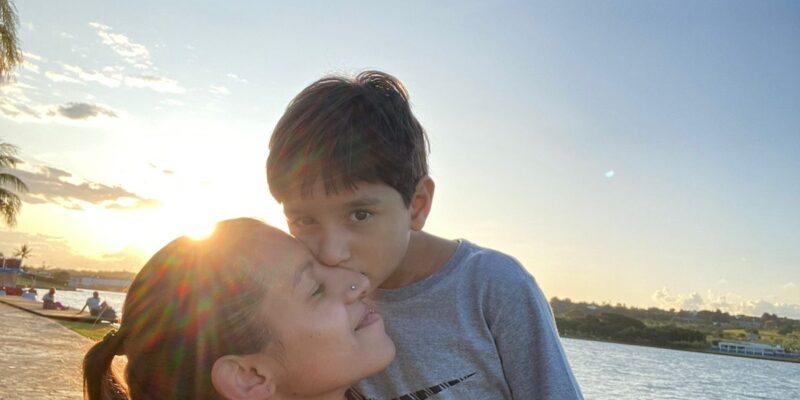 Mulher com cabelo preso recebe beijo de uma criança no rosto, em momento de afeto. Ao fundo, há um lago, gramado e calçada, com o sol baixo no horizonte iluminando a cena.