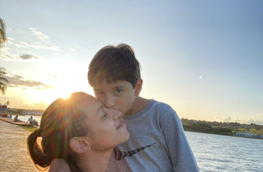Mulher com cabelo preso recebe beijo de uma criança no rosto, em momento de afeto. Ao fundo, há um lago, gramado e calçada, com o sol baixo no horizonte iluminando a cena.