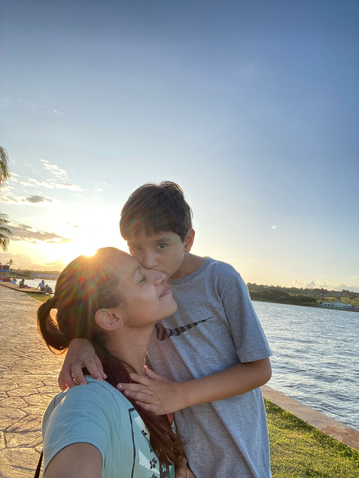 Mulher com cabelo preso recebe beijo de uma criança no rosto, em momento de afeto. Ao fundo, há um lago, gramado e calçada, com o sol baixo no horizonte iluminando a cena.