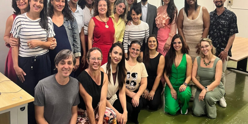 Grupo de homens e mulheres posa em sala de reunião, sorrindo. Parte está em pé e outra sentada. Ambiente interno com mesa, quadro ao fundo e clima de integração e trabalho coletivo.