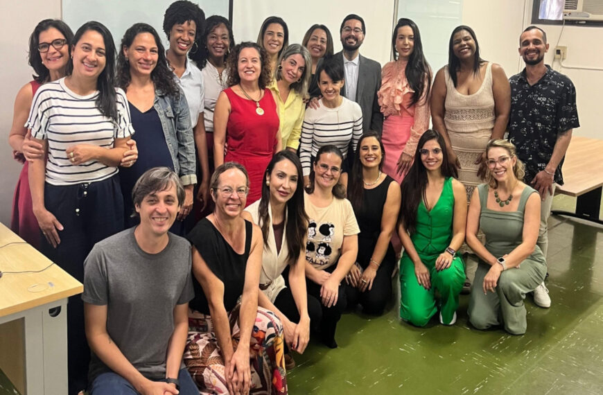 Grupo de homens e mulheres posa em sala de reunião, sorrindo. Parte está em pé e outra sentada. Ambiente interno com mesa, quadro ao fundo e clima de integração e trabalho coletivo.