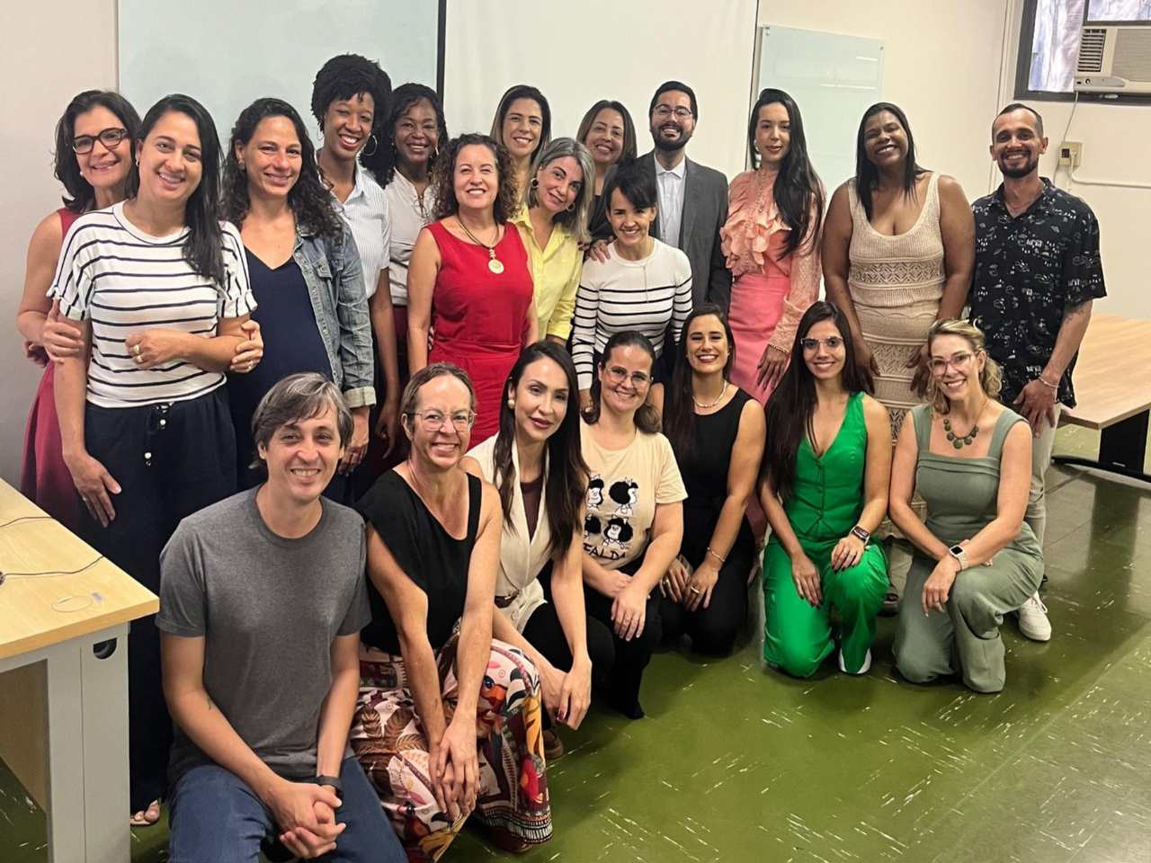 Grupo de homens e mulheres posa em sala de reunião, sorrindo. Parte está em pé e outra sentada. Ambiente interno com mesa, quadro ao fundo e clima de integração e trabalho coletivo.