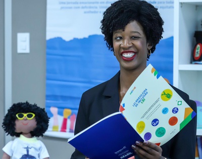A secretária nacional Isadora Nascimento sorri enquanto segura material da Lei Brasileira de Inclusão (LBI), com capa branca e elementos em azul, verde, amarelo e vermelho. Ao fundo, há banner azul do Novo Viver sem Limite e, ao lado, uma boneca com óculos de sol amarelos e lentes pretas.