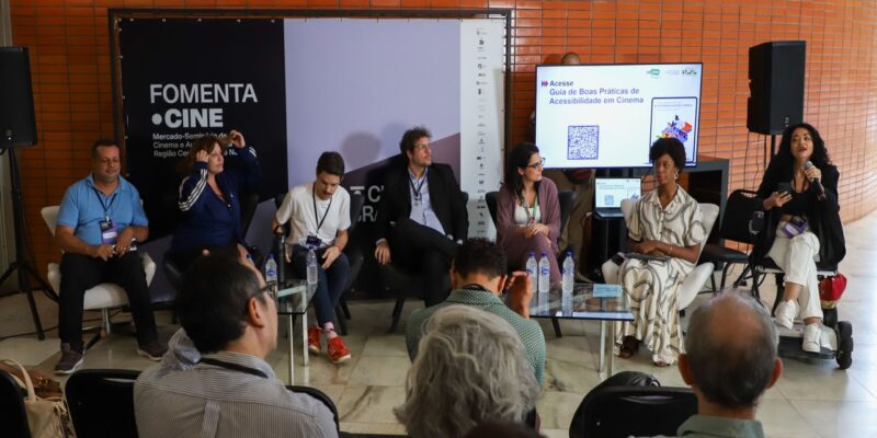 Painel do FomentaCine com sete participantes sentados em poltronas, em debate sobre acessibilidade no cinema. Ao fundo, banner do evento e tela com QR code do guia; público acompanha sentado à frente.