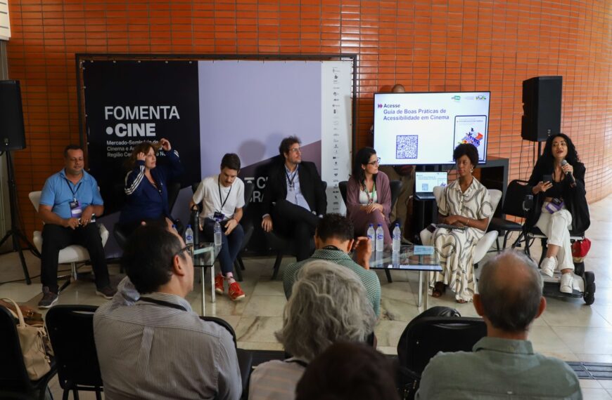 Painel do FomentaCine com sete participantes sentados em poltronas, em debate sobre acessibilidade no cinema. Ao fundo, banner do evento e tela com QR code do guia; público acompanha sentado à frente.