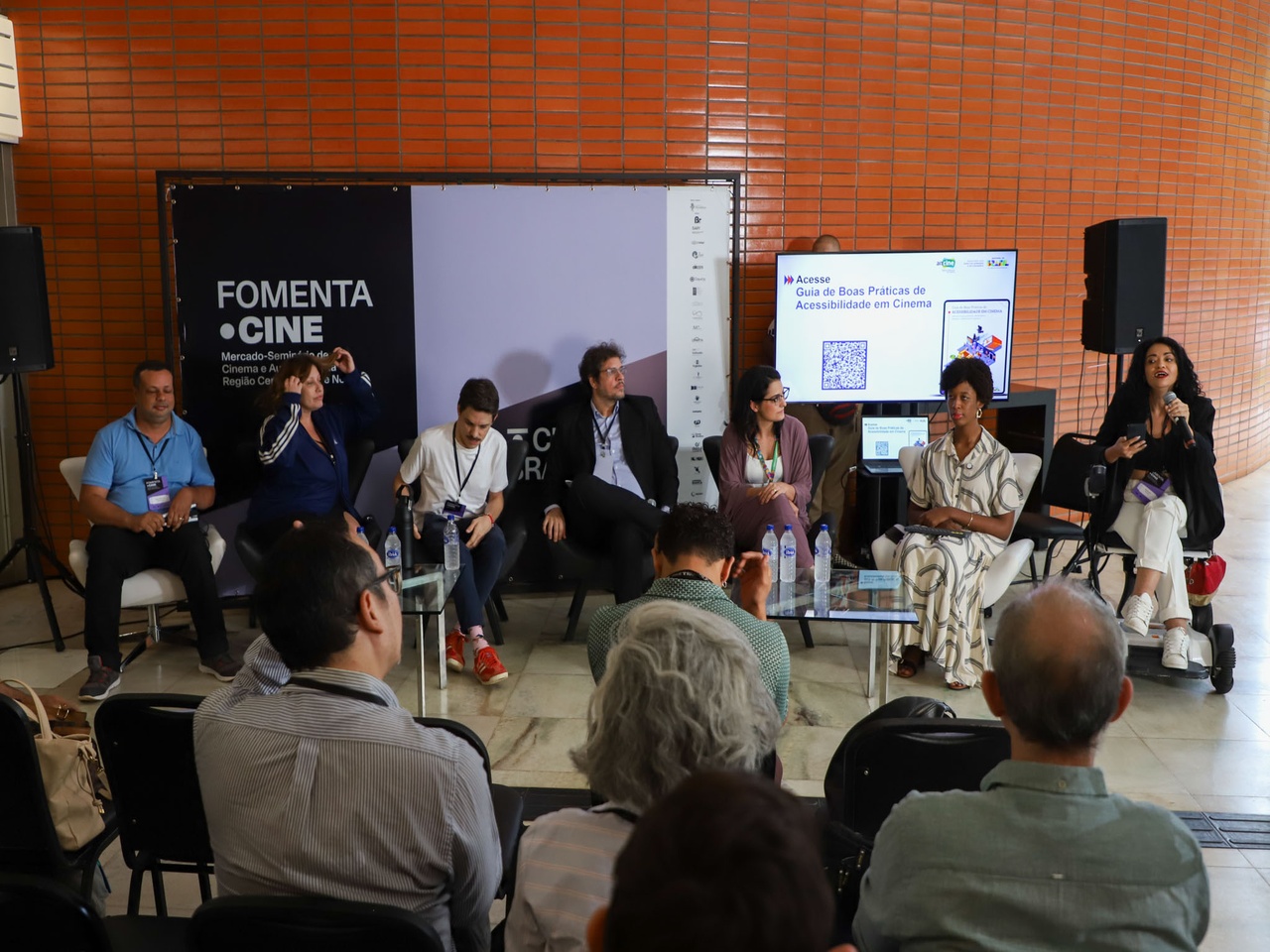 Painel do FomentaCine com sete participantes sentados em poltronas, em debate sobre acessibilidade no cinema. Ao fundo, banner do evento e tela com QR code do guia; público acompanha sentado à frente.