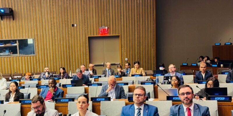 Auditório institucional com participantes sentados em fileiras, usando crachás e acompanhando a atividade. Pessoas de diferentes perfis observam atentos, com laptops e microfones sobre as mesas. Ambiente formal.