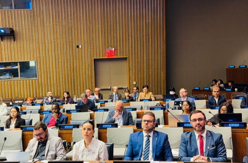 Auditório institucional com participantes sentados em fileiras, usando crachás e acompanhando a atividade. Pessoas de diferentes perfis observam atentos, com laptops e microfones sobre as mesas. Ambiente formal.