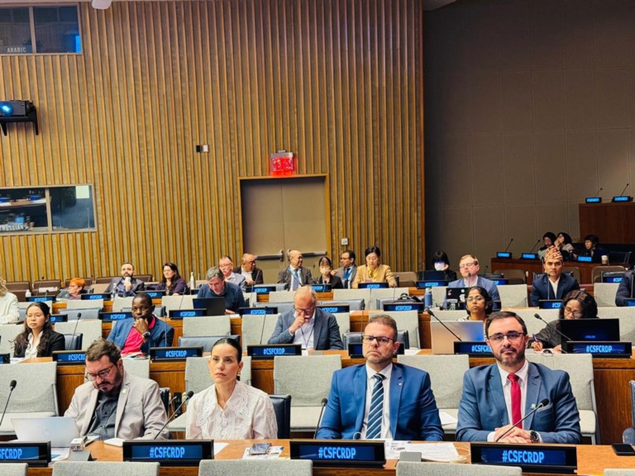 Auditório institucional com participantes sentados em fileiras, usando crachás e acompanhando a atividade. Pessoas de diferentes perfis observam atentos, com laptops e microfones sobre as mesas. Ambiente formal.
