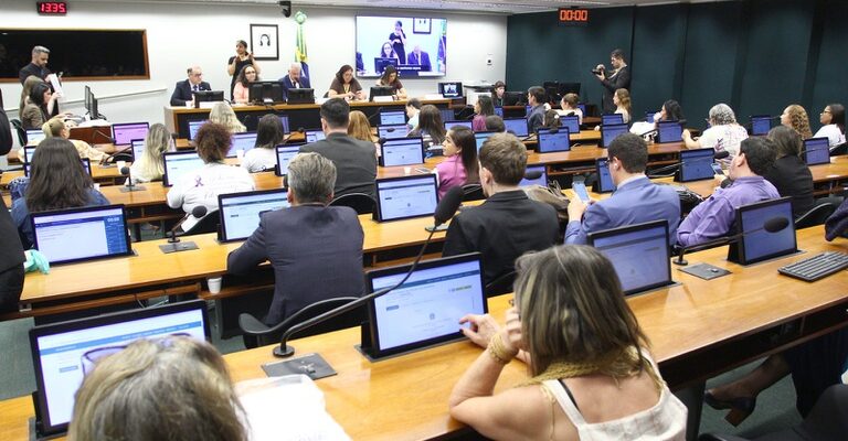 A imagem mostra uma reunião ou audiência pública em um plenário de comissões da Câmara dos Deputados, com participantes sentados em bancadas equipadas com computadores e microfones, voltados para uma mesa diretora ao fundo. O ambiente é formal, com presença da bandeira do Brasil e painéis eletrônicos exibindo a transmissão do evento e uma intérprete de Libras.