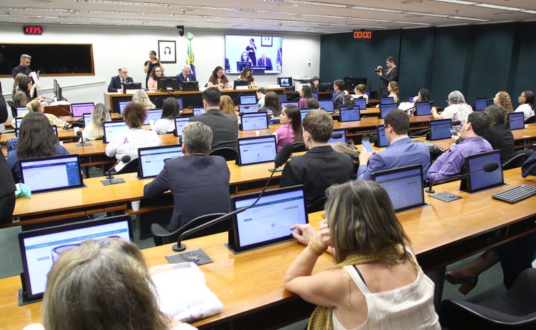 A imagem mostra uma reunião ou audiência pública em um plenário de comissões da Câmara dos Deputados, com participantes sentados em bancadas equipadas com computadores e microfones, voltados para uma mesa diretora ao fundo. O ambiente é formal, com presença da bandeira do Brasil e painéis eletrônicos exibindo a transmissão do evento e uma intérprete de Libras.