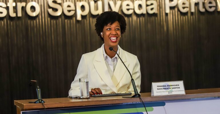 Uma mulher negra está sentada atrás de uma mesa de madeira em um auditório institucional. Ela usa um blazer branco e sorri enquanto fala ao microfone. À frente dela, há um copo com água, um notebook fechado e uma placa de identificação com o nome “Isadora Rodrigues Nascimento Santos”. Ao fundo, uma parede escura com letras grandes em relevo compõe o cenário do evento. A iluminação destaca a participante no centro da imagem, transmitindo um ambiente formal e acolhedor.