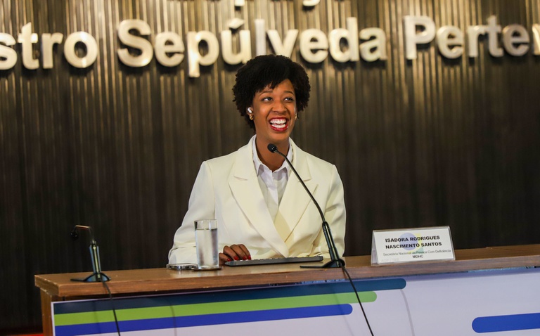 Uma mulher negra está sentada atrás de uma mesa de madeira em um auditório institucional. Ela usa um blazer branco e sorri enquanto fala ao microfone. À frente dela, há um copo com água, um notebook fechado e uma placa de identificação com o nome “Isadora Rodrigues Nascimento Santos”. Ao fundo, uma parede escura com letras grandes em relevo compõe o cenário do evento. A iluminação destaca a participante no centro da imagem, transmitindo um ambiente formal e acolhedor.